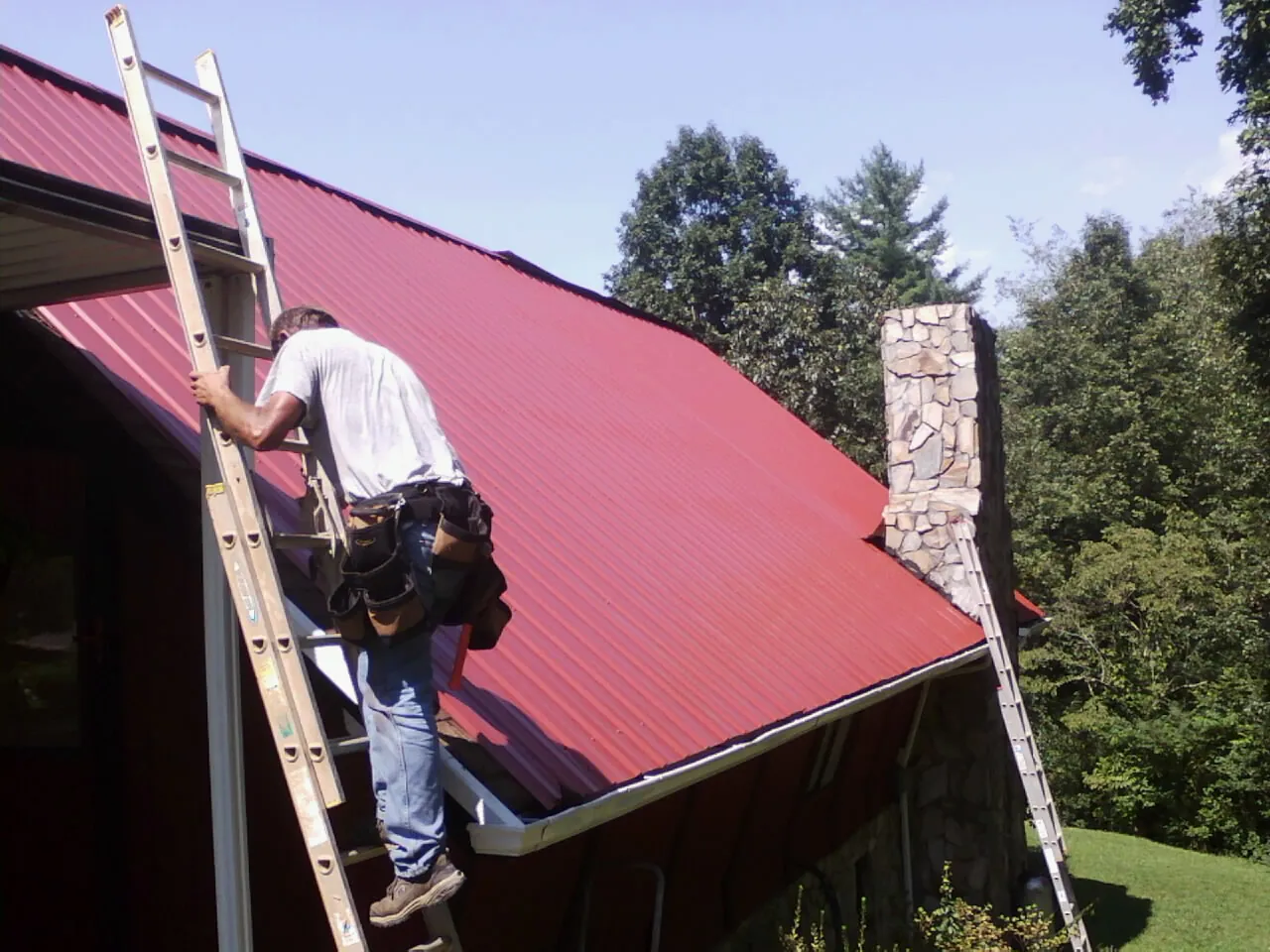 Roofer on a ladder beside a red metal roof and stone chimney — Soffit & Fascia Repair by Roofing Brothers in Sheyenne